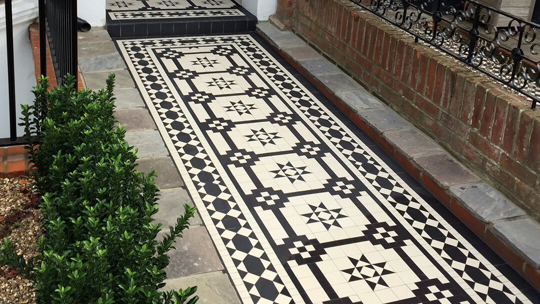 Black and White geometric mosaic path tiles framed by stone slabs. Decorative iron railings sit on top of a small red brick wall.