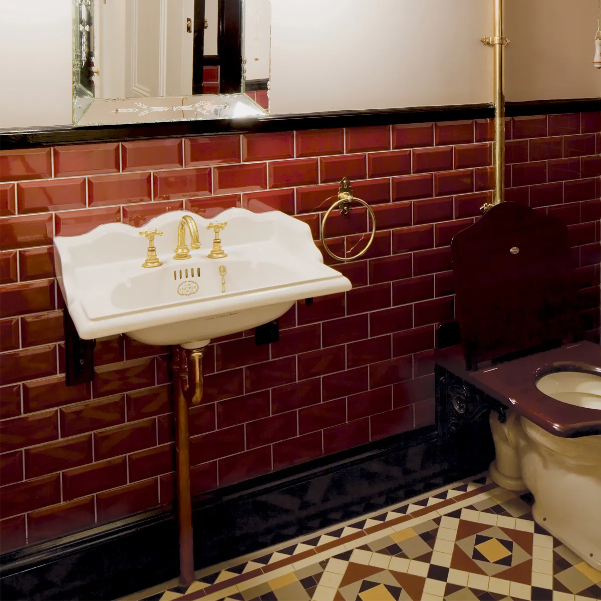 Deep red metro tiles with black dado rail behind Victorian toilet and sink with brass fittings. On the floor a traditional Victorian mosaic tile design.