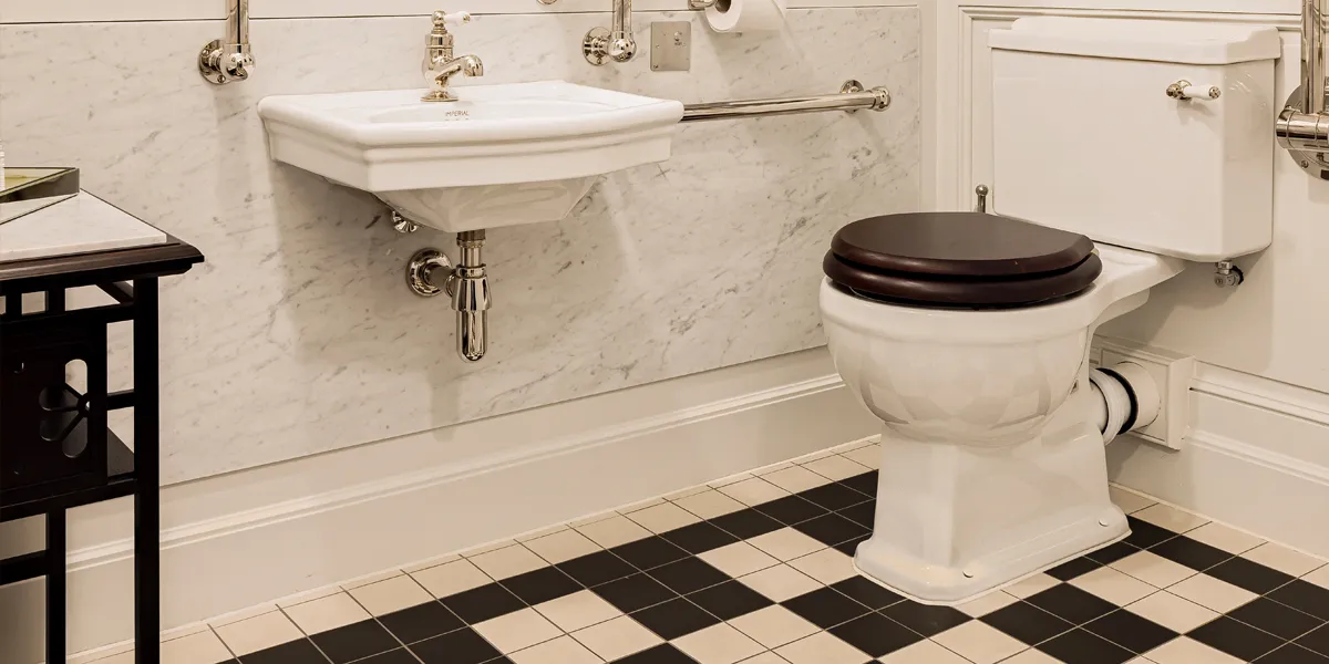 Larger format black and white tiles on the floor of a bathroom with Edwardian style sink and toilet. Dark wood vanity desk and toilet seat contrast against the white marble and wooden panelling on the wall.